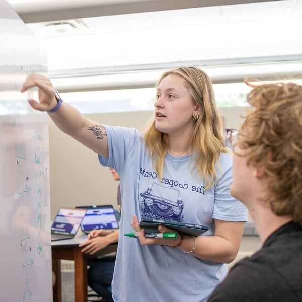 A female student standing in front of a whiteboard point to a formula as a seated male student looks on.